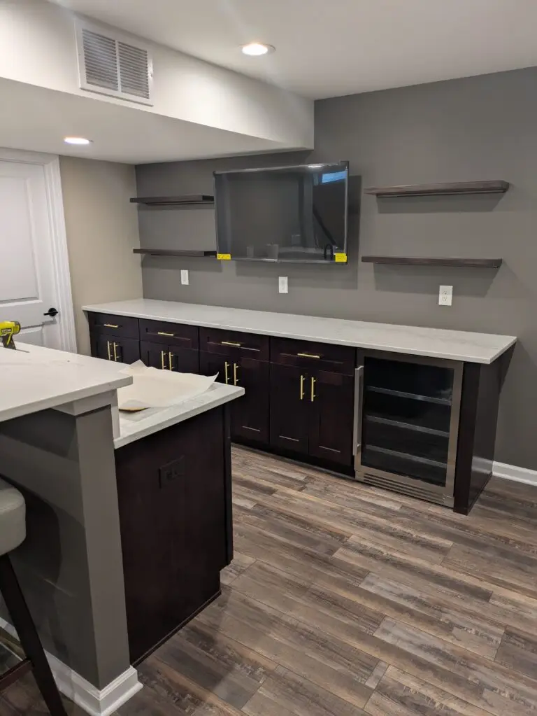 Finished basement wet bar with dark cabinets, wine fridge, floating shelves and wall-mounted TV